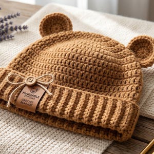 Puede incluir: Un gorro de ganchillo color canela con dos orejas de oso redondeadas en la parte superior y un borde acanalado. Una pequeña etiqueta rectangular marrón está sujeta al frente con un lazo de cuerda y un botón, mostrando el texto HUGGABLE STITCHES CROCHET BEAR HAT.