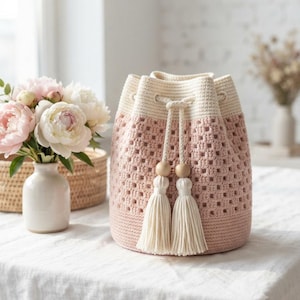 May include: A crochet bucket bag featuring a dusty pink body with a square-patterned texture and a solid cream-coloured top section. The bag includes a cream drawstring closure finished with two wooden beads and long, matching cream-coloured tassels.