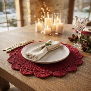 May include: A round, deep red crocheted placemat with a scalloped edge, featuring a white ceramic plate topped with a folded cream-colored napkin tied with twine and a small sprig of evergreen, set on a wooden table with gold cutlery.