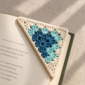 May include: A triangular crochet bookmark featuring a granny square pattern in cream, light blue, and dark blue yarn. The bookmark is placed on the corner of an open book titled The Shadow of the Wind, with visible printed text on the page.