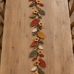 Può includere: Una lunga ghirlanda decorativa realizzata in filo all'uncinetto, caratterizzata da un motivo ripetitivo di piccoli fiori gialli e foglie variegate nei toni dell'arancio ruggine, verde scuro e crema, unite da un sottile viticcio marrone.