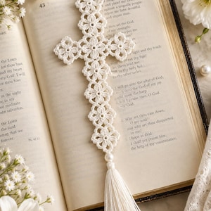 May include: A white crochet bookmark shaped like a cross, constructed from a series of interconnected floral motifs. The bookmark features a single white pearl bead at the base, which connects to a long, straight white thread tassel.