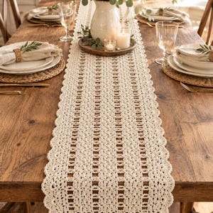 May include: A long, cream-colored crochet table runner featuring an intricate openwork pattern with vertical columns and a scalloped edge. The runner is crafted from yarn and displays a detailed, repeating geometric design that runs the full length.