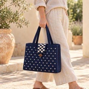 May include: A rectangular navy blue crochet tote bag featuring a repeating pattern of small white dots. The bag has two matching navy blue handles and a central flap closure with a checkered white and navy pattern secured by a brown button.