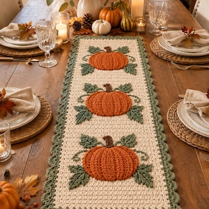 May include: A rectangular crochet table runner featuring an off-white base with three centered orange pumpkins, each surrounded by green leaves and vines. The runner has a scalloped green border and is displayed on a wooden dining table.