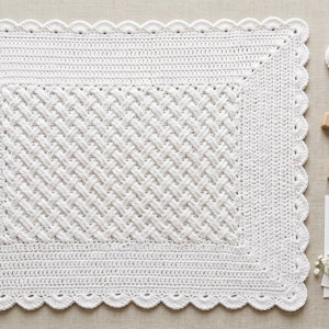 May include: A rectangular white crochet blanket with a central basketweave pattern and a scalloped border. Beside it are two white crochet booties, a wooden cross, a rolled ribbon, and a card with the text Blessing Day Celebrating Eliza Jane.