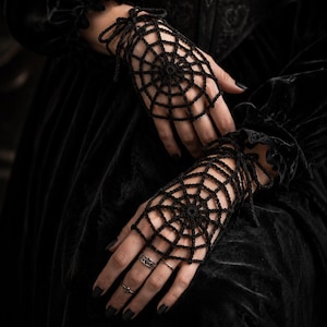May include: A pair of black fingerless gloves featuring a crocheted spider web pattern. The gloves are designed to cover the back of the hand and are secured at the wrist with thin, matching black ties. The intricate web design radiates from a central point.