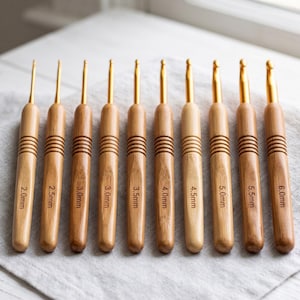 Può includere: Un set di dieci uncinetti con manici in legno marrone chiaro e punte in metallo dorato. Ogni manico presenta quattro scanalature orizzontali ed è inciso al laser con misure che vanno da 2,0 mm a 6,0 mm con incrementi di 0,5 mm.