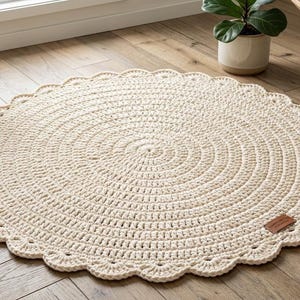 May include: A round, cream-colored crocheted rug featuring a textured, concentric circular pattern and a decorative scalloped edge. A small, rectangular brown leather-like brand tag is attached to the outer edge of the rug.