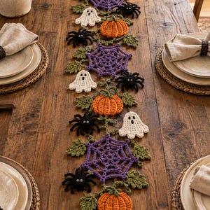 May include: A Halloween-themed crochet table runner featuring orange pumpkins, purple spiderwebs, black spiders, and white ghosts connected by green leafy vines. The decorative runner is arranged in a vertical line down the center of a wooden table.