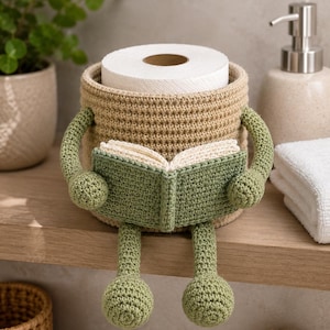 May include: A beige crochet toilet paper holder featuring a character with sage green arms and legs. The character is holding an open book with cream-colored pages and a sage green cover. The holder is cylindrical and designed to fit a standard roll.