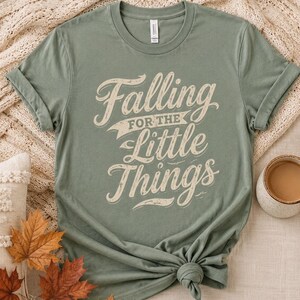 May include: A sage green short-sleeved t-shirt featuring the cream-coloured text "Falling FOR THE Little Things" in a stylised, distressed font. The shirt has a crew neckline and is shown with the sleeves rolled up and a knot at the bottom hem.