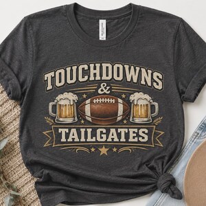 Könnte beinhalten: Ein dunkelgraues Kurzarm-T-Shirt mit einem Grafikdruck und dem Text "TOUCHDOWNS & TAILGATES". Das Design zeigt einen braunen Football, der zwischen zwei mit Bier gefüllten Glaskrügen zentriert ist, akzentuiert durch Sterne und dekorative Banner.