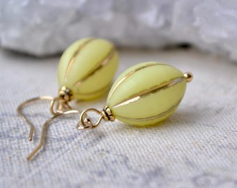 May include: A pair of pale yellow oval-shaped earrings featuring vertical gold-toned metallic stripes. Each earring is suspended from a gold-toned metal hook and includes a small gold-toned metal bead at the top of the bead-shaped component.