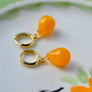 May include: A pair of gold-toned hoop earrings, each featuring a smooth, vibrant orange teardrop-shaped pendant. The small, circular hoops are polished and metallic, with the bright orange drops suspended from a simple gold-coloured jump ring.