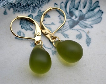 May include: A pair of gold-tone leverback earrings featuring smooth, teardrop-shaped olive green glass beads. The earrings have a polished metallic finish on the hooks and a matte, translucent appearance on the rounded, drop-style glass components.