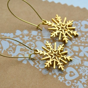 May include: A pair of gold-coloured snowflake earrings. Each earring features a detailed snowflake design with a star in the centre. The earrings hang from a simple gold-coloured wire hook. The background is a brown surface with a white pattern.