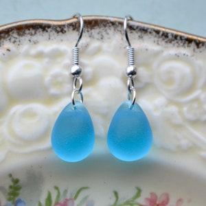 The image shows a pair of teardrop earrings with bright blue -almost turquoise - glass teardrops suspended from silver-plated fishhook earring wires. The glass beads have been etched to give them a sea glass appearance.