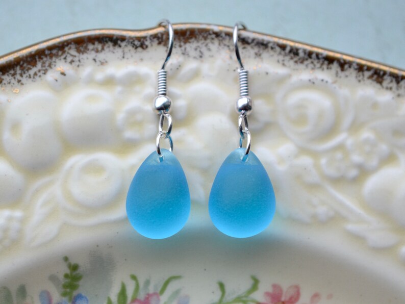 The image shows a pair of teardrop earrings with bright blue -almost turquoise - glass teardrops suspended from silver-plated fishhook earring wires. The glass beads have been etched to give them a sea glass appearance.