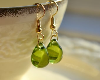 May include: A pair of gold-colored drop earrings with pear-shaped, translucent green glass beads. The earrings hang from gold-colored hooks and are displayed against a blurred background.