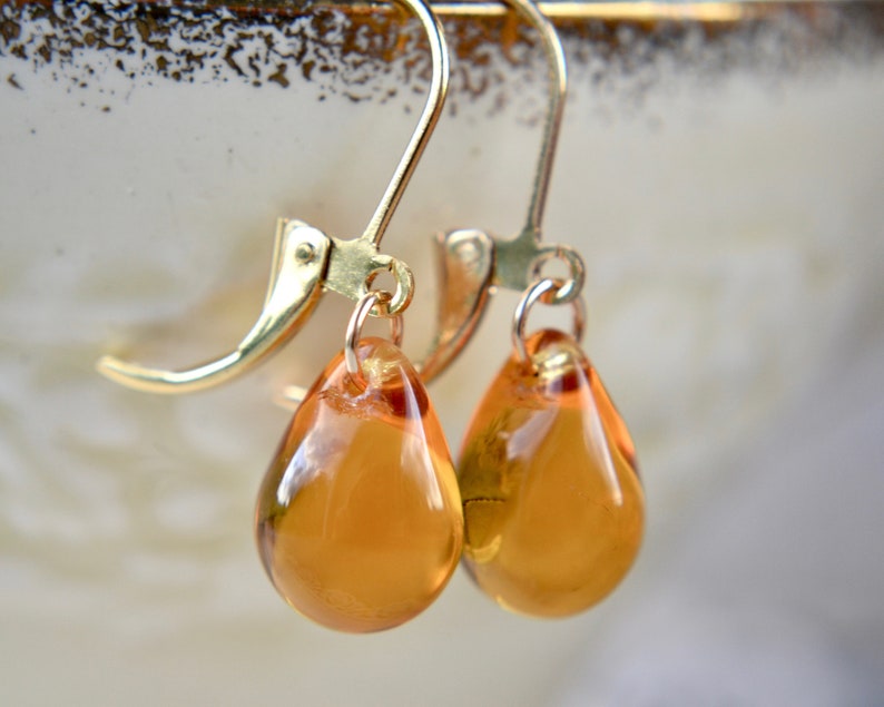 This image shows golden yellow glass teardrops suspended from gold-plated leverback hooks. They are suspended from the rim of a vintage teacup.