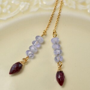 May include: Elegant gold earrings with hanging chains. Each chain has pale blue beads and a deep red, teardrop-shaped gemstone. The earrings are displayed on a cream-coloured surface.