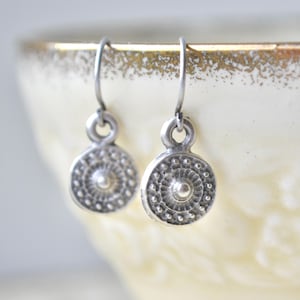 Peut inclure: Une paire de boucles d'oreilles en argent avec un motif circulaire texturé. Chaque boucle d'oreille présente un petit point central surélevé entouré de cercles concentriques et de petits points. Les boucles d'oreilles sont suspendues à de simples crochets incurvés.