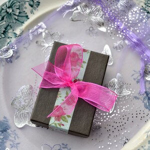 Puede incluir: Una peque&ntilde;a caja de regalo marr&oacute;n con una cinta rosa y papel con estampado floral. La caja est&aacute; sobre una tela morada y blanca con dise&ntilde;os de mariposas plateadas.