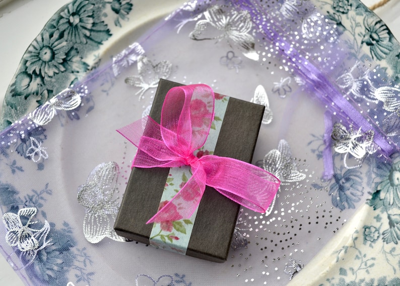 Puede incluir: Una peque&ntilde;a caja de regalo marr&oacute;n con una cinta rosa y papel con estampado floral. La caja est&aacute; sobre una tela morada y blanca con dise&ntilde;os de mariposas plateadas.