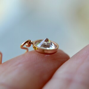 Puede incluir: Primer plano de un colgante dorado con una piedra preciosa transparente y facetada. El colgante se sostiene en una mano.