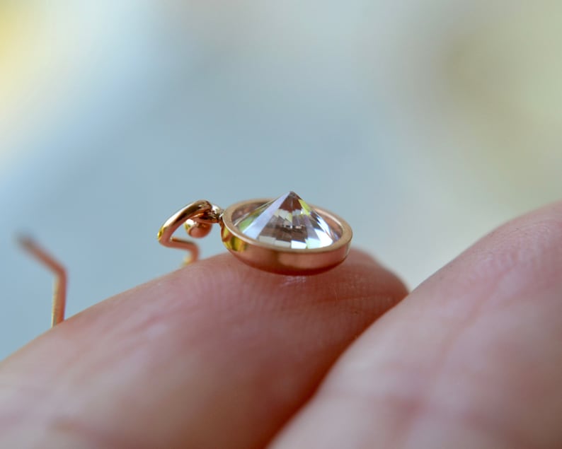 Puede incluir: Primer plano de un colgante dorado con una piedra preciosa transparente y facetada. El colgante se sostiene en una mano.