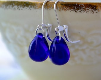 May include: A pair of drop earrings featuring smooth, teardrop-shaped cobalt blue glass beads suspended from polished silver-tone leverback hooks. The vibrant blue glass has a translucent quality, catching the light to highlight the rounded shape.
