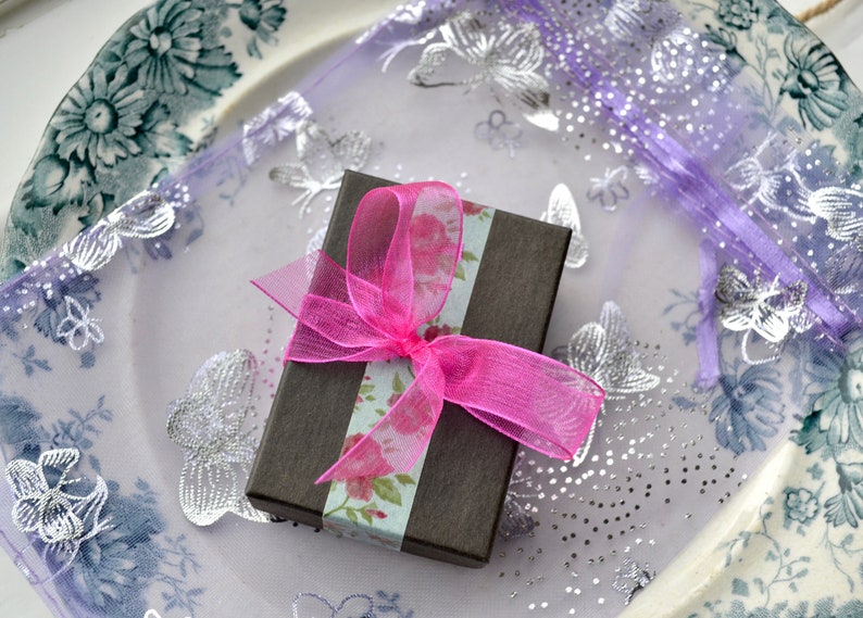 May include: A small, brown gift box with a pink ribbon and floral patterned paper. The box is sitting on a purple and white fabric with silver butterfly designs.