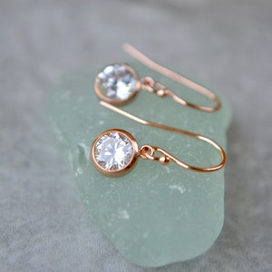 Puede incluir: Un par de pendientes colgantes de oro rosa con piedras redondas transparentes. Los pendientes est&aacute;n sobre una superficie verde claro.