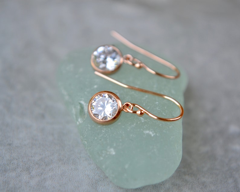 Puede incluir: Un par de pendientes colgantes de oro rosa con piedras redondas transparentes. Los pendientes est&aacute;n sobre una superficie verde claro.