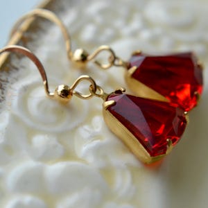 May include: A pair of gold-toned earrings featuring faceted, deep red gemstones. The earrings have a classic hook design and are set in a gold-colored metal. The gemstones are a vibrant, eye-catching color.