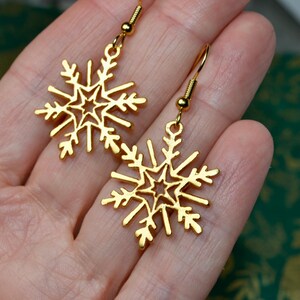 以下が含まれることがあります： 金色の雪の結晶のイヤリングのペア。各イヤリングは、中央に星が付いた詳細な雪の結晶のデザインが特徴です。イヤリングは金色のフックから吊り下げられており、冬や休日に最適です。