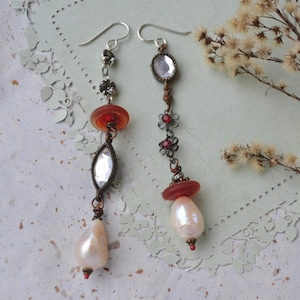 May include: Dangling earrings with a vintage aesthetic. Each earring features a silver hook, a cluster of small black beads, a round orange bead, and a clear, oval-shaped stone. A teardrop-shaped pearl hangs at the bottom. The earrings are set against a textured background.