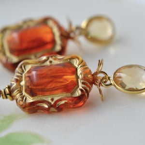 May include: Close-up of a pair of ornate earrings. Each earring features a rectangular, amber-coloured stone set in a gold-toned frame. A smaller, round, pale yellow stone dangles below.