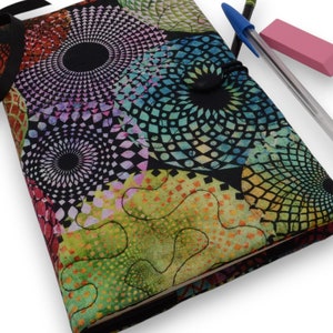 May include: A colorful, quilted journal with a black ribbon closure. The cover features a vibrant pattern of geometric shapes in shades of green, blue, orange, and black. A blue pen and pink eraser rest on the journal.