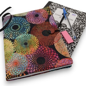 May include: A black notebook with a colorful mandala pattern cover. The notebook is closed and has a black ribbon tied around it. A composition book, pen, and pink eraser are tucked into the notebook.