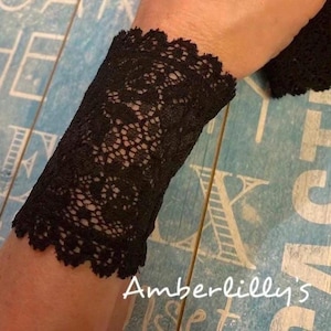 May include: Black lace wrist cuff with a scalloped edge. The cuff is made of delicate lace and has a floral pattern.