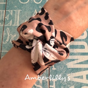 Peut inclure: Un bracelet de poignet en tissu noué avec un motif animalier beige et noir. Le bracelet est enroulé autour d'un poignet. Le texte "Amberlilly's" est visible en écriture blanche.