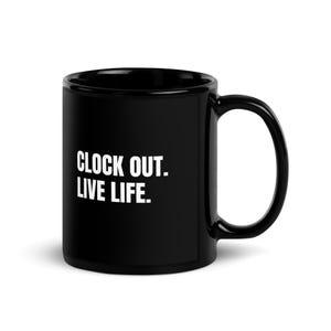 May include: A black ceramic mug featuring the white text CLOCK OUT. LIVE LIFE. printed on the front. The mug has a smooth, glossy finish, a cylindrical body, and a curved handle on the right side, providing a simple and functional design.