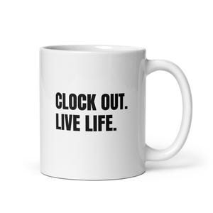 May include: A white ceramic mug with a curved handle featuring the black text CLOCK OUT. LIVE LIFE. printed in a bold, sans-serif font on the front. The mug has a smooth, glossy finish and a standard cylindrical shape with a flat base.