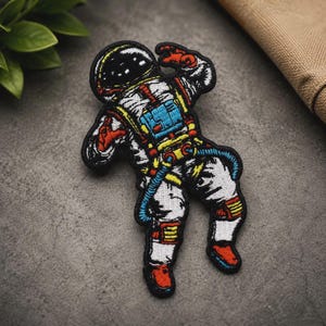 May include: An embroidered patch featuring an astronaut in a white spacesuit with black, blue, yellow, and red accents. The astronaut is depicted in a floating pose with a black helmet visor, a blue chest pack, and red gloves and boots.