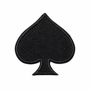 May include: A solid black spade suit symbol patch featuring a dense, textured embroidery pattern with a clearly defined stitched border edge. The shape is symmetrical, representing the classic playing card icon with a pointed top and stem base.
