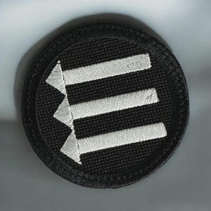 May include: A circular embroidered patch featuring a black background with three horizontal white arrows pointing to the left. The design is stitched with a thick black border, creating a clean and defined edge for this textile accessory.