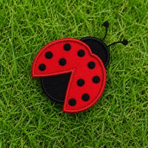 May include: A ladybug-shaped embroidered patch featuring a bright red wing section with black circular spots and a solid black head section. The design includes two thin black antennae extending from the top of the head, with a stitched border.
