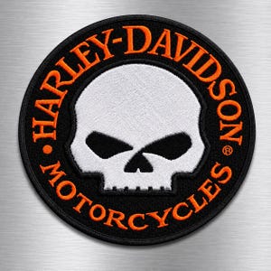 May include: A circular embroidered patch featuring a white skull graphic centered on a black background. The text HARLEY-DAVIDSON MOTORCYCLES is stitched in orange thread around the skull, with a registered trademark symbol on the right side.